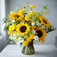 Bouquet of sunflowers, yellow daisies, and white roses in a clear vase on a wooden surface with a light gray background.